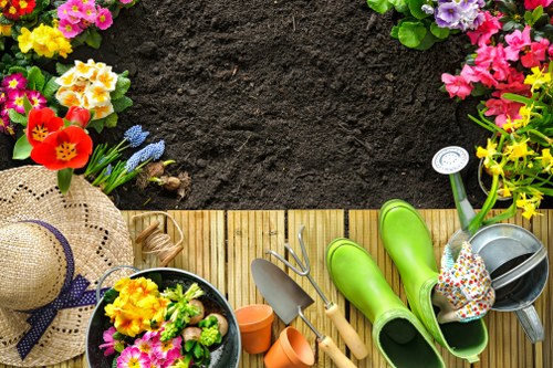 The image shows a well-maintained garden space with a neatly prepared wooden deck bordered by colourful flowering plants, including pink, purple, yellow, and red blooms, creating a vibrant and inviting border. Directly adjacent to the deck is a patch of rich, dark soil, suggesting recent planting or preparation for landscaping work. On the deck, various gardening tools and accessories are visible, including bright green rubber gardening boots, a metal watering can, small terracotta pots, and a pair of pruning shears, indicating active outdoor maintenance. A straw sunhat with a navy blue ribbon is placed on the edge of the deck, adding a touch of seaside charm. The lawn area in the foreground is lush and green, with a dense, healthy grass texture, contrasting with the dark soil and providing a soft ground cover. The overall scene depicts an outdoor space in good condition, with signs of ongoing gardening activity, set in natural outdoor lighting under a clear sky, supporting professional gardening and landscaping services in Queens Park or nearby areas.