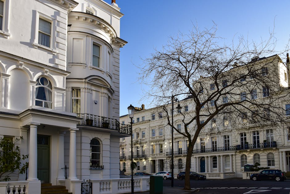 A white Victorian-style terrace house with ornate architectural details, including decorative moldings and columns at the entrance, situated in Queens Park. In the foreground, a leafless tree with a g
